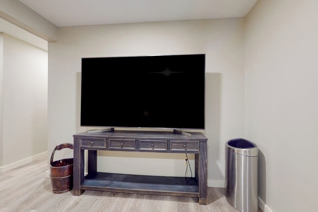 A room with a flat-screen TV mounted on the wall, a wooden console table with drawers, a trash can, and a wooden bucket.