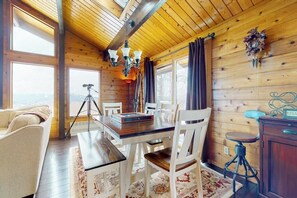 Ein Holzblockhaus-Interieur mit einem Esstisch, weißen Stühlen, einer Chandelüster und einem Stativ in der Ecke.