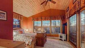 Une chambre avec un plafond en bois, un lit recouvert d’une couvre-lit à motifs floraux, une table de chevet, une commode et de grandes fenêtres avec des rideaux.