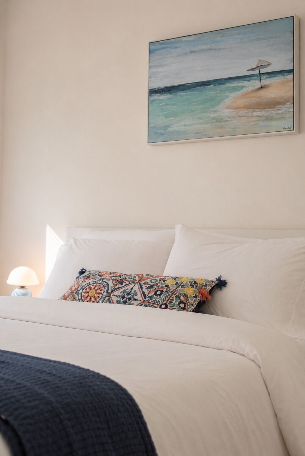 A bedroom with a bed, a colorful pillow, a lamp, and a painting of a beach scene.