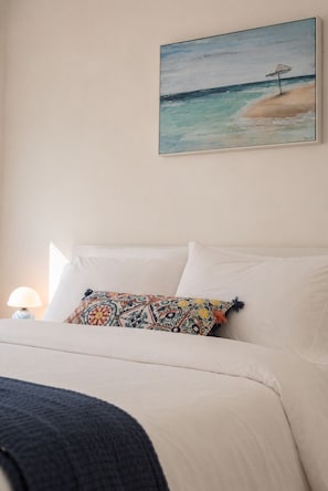 Ein Schlafzimmer mit einem Bett, einem farbenfrohen Kissen, einer Lampe und einem Bild einer Strandlandschaft.