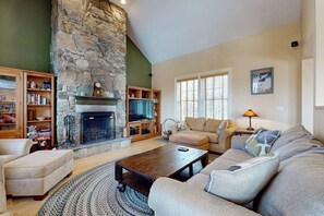 A living room with a stone fireplace, a television, a sofa, and a coffee table. - Beautiful house w/ mountain & pond views, hot tub, pool table, bbq, firepit, w/d (Windham)