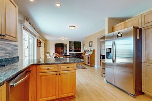 A kitchen with wooden cabinets, a granite countertop, a stainless steel refrigerator, and a dining area with a table and chairs. - Beautiful house w/ mountain & pond views, hot tub, pool table, bbq, firepit, w/d (Windham)