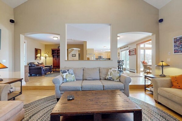 A spacious living room with a large sofa, a coffee table, and a kitchen in the background. - Windham home with pond views, hot tub, sunroom and pool table (Windham)