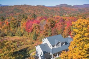A house with a sloped roof, surrounded by a forest with autumn foliage. - Beautiful house w/ mountain & pond views, hot tub, pool table, bbq, firepit, w/d (Windham)