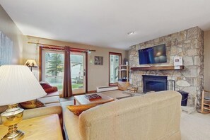 A living room with a stone fireplace, a flat-screen TV mounted on the wall, a sofa, a coffee table, and a lamp. - Ski-in/ski-out condo w/ deck, wood-burning fireplace, wifi, community amenities (West Dover)