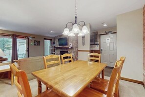 A dining area with a wooden table and chairs, a hanging light fixture, a stone fireplace, and a television mounted on the wall. - Ski-in/ski-out condo w/ deck, wood-burning fireplace, wifi, community amenities (West Dover)