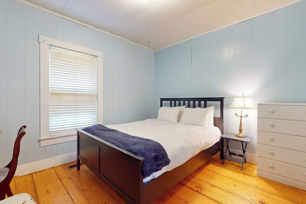 Ein Schlafzimmer mit einem Bett, einem Schminktisch, einem Nachttisch mit Lampe, einem Fenster mit Jalousien und einem Sessel.