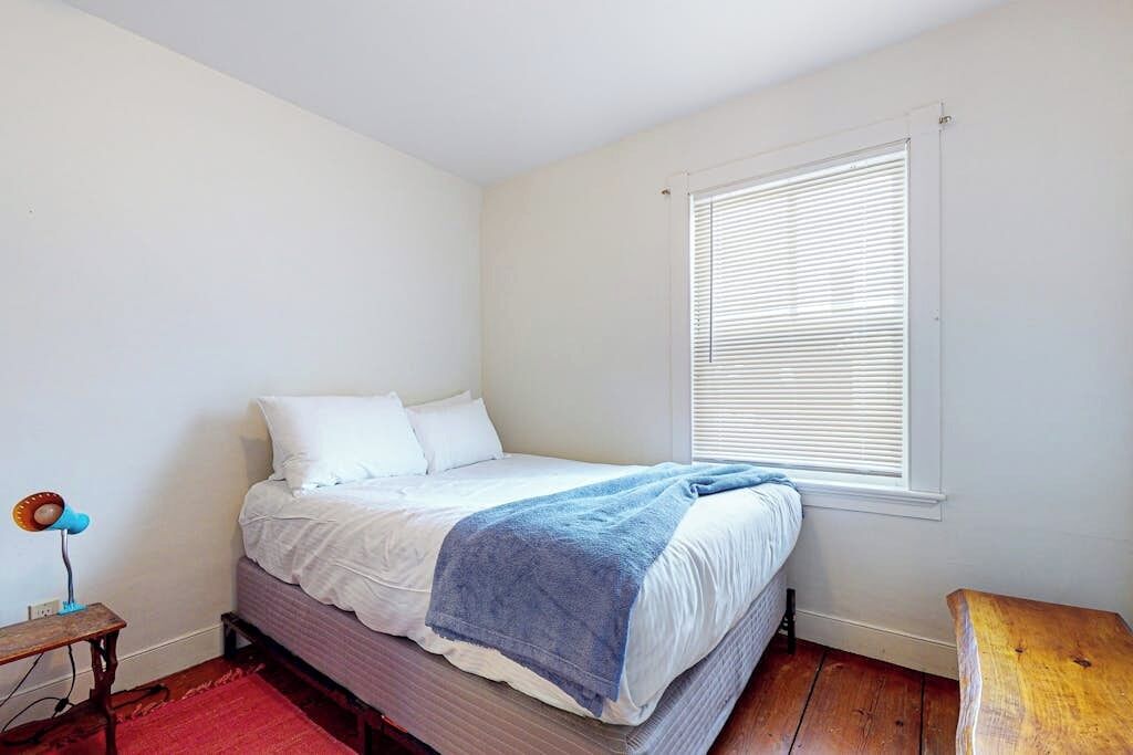 A bedroom with a bed, a window with blinds, a red rug, and a wooden bench.
