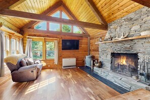 A log cabin interior with a stone fireplace, wooden floors, a large window, and a cozy seating area.