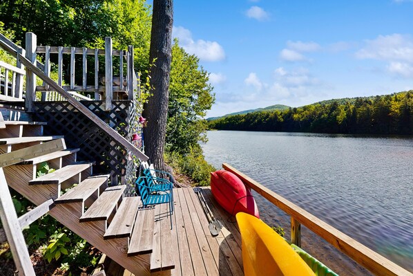 A wooden deck with a staircase leading to a deck chair and a kayak, overlooking a calm lake with a forested shoreline.