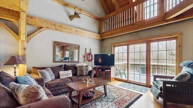 A cozy living room with a wooden ceiling, a large window, a television, and a sofa with a cushion labeled 'COZY'. - Spacious townhouse w/ gas fireplace, balcony, game room,  w/d,  loft (Dover)