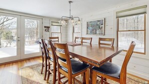 A dining room with a wooden table and chairs, a chandelier, and large windows with a view of trees. - Cozy farmhouse w/ mountain views, woodstove, bbq, cable, close to Okemo, w/d (Cavendish)