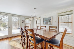 A dining room with a wooden table and chairs, a chandelier, and large windows with a view of trees. - Cozy farmhouse w/ mountain views, woodstove, bbq, cable, close to Okemo, w/d (Cavendish)