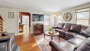A living room with a brown leather sofa, a round coffee table, and a large clock on the wall. - Cozy farmhouse w/ mountain views, woodstove, bbq, cable, close to Okemo, w/d (Cavendish)