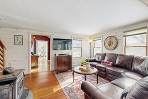 A living room with a brown leather sofa, a round coffee table, and a large clock on the wall. - Cozy farmhouse w/ mountain views, woodstove, bbq, cable, close to Okemo, w/d (Cavendish)