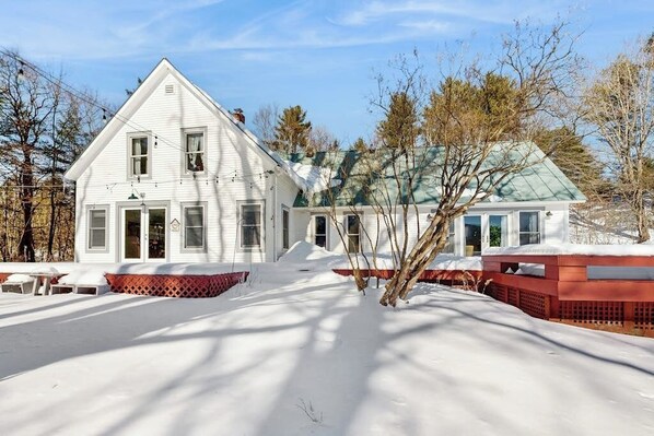 A white house with a green roof, surrounded by snow-covered ground and bare trees. - Cozy farmhouse w/ mountain views, woodstove, bbq, cable, close to Okemo, w/d (Cavendish)