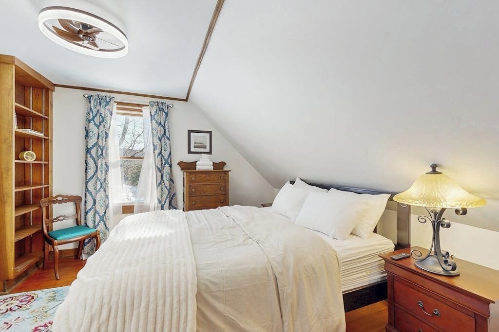 Un dormitorio con cama, una mesita de noche con lámpara, una silla de madera, una estantería y una ventana con cortinas.