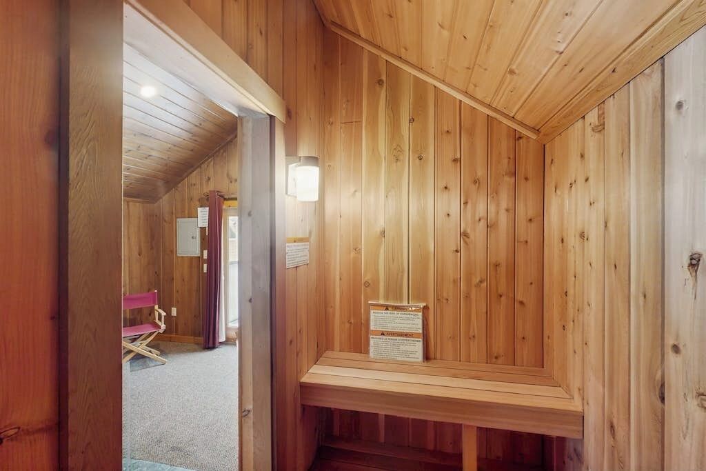 A wooden sauna room with a bench, a wall-mounted light, and a door leading to another room.
