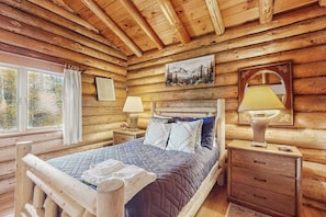 Ein Schlafzimmer in einer Blockhütte mit einem Bett, Nachttischen, einem Fenster mit Vorhängen, einem Gemälde und einem Spiegel.