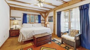 A bedroom with a bed, wooden beams, a ceiling fan, a chair, a window with curtains, and a rug. - Lakefront house w/ mountain views, waterfront, gameroom, gym, a/c, w/d (Ludlow)