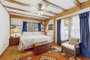 Ein Schlafzimmer mit Bett, Holzbalken, Deckenventilator, Stuhl, Fenster mit Vorhängen und Teppich.