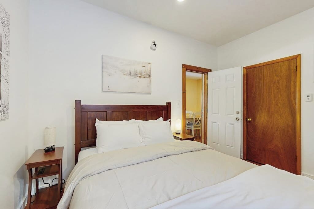 Un dormitorio con una cama de madera, una mesita de noche con lámpara, una puerta y un reloj en la pared.