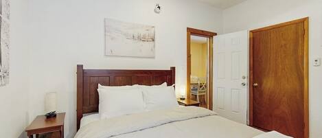 Un dormitorio con una cama de madera, una mesita de noche con lámpara, una puerta y un reloj en la pared.