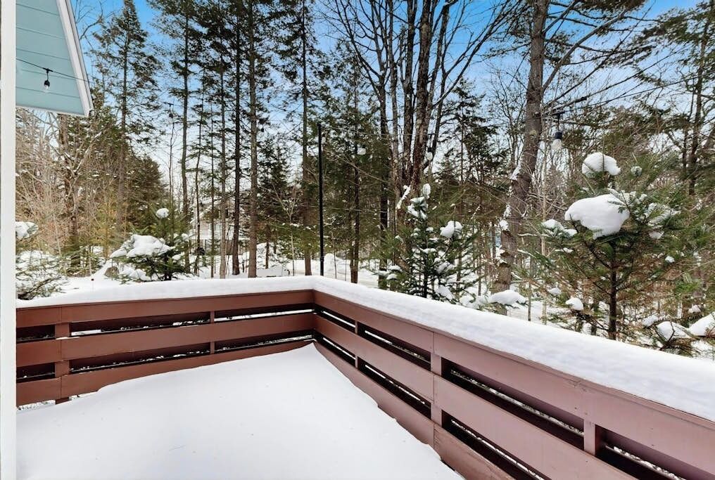 A snow-covered deck with a wooden railing, surrounded by tall trees and a clear sky.