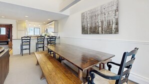 Un comedor con una larga mesa de madera, cuatro sillas y un banco. Se observa una cocina al fondo con armarios blancos y un refrigerador.