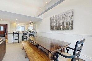 Ein Essbereich mit einem langen Holztisch, vier Stühlen und einer Bank. Im Hintergrund befindet sich eine Küche mit weißen Schränken und einem Kühlschrank.