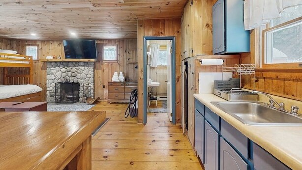 A cabin interior with a stone fireplace, wooden floors, a kitchen with a sink, and a television mounted on the wall. - Cozy studio cabin w/ seasonal mountain views, wrap-around deck, window a/c, bbq (Wardsboro)