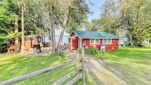 A red cabin with a green roof, surrounded by trees and a grassy area. - Dog-Friendly Lakefront Home with WiFi, Private Dock & Firepit (Grand Isle)
