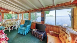 A cabin with a dining table, chairs, a television on a chest, and a large window overlooking a lake. - Dog-Friendly Lakefront Home with WiFi, Private Dock & Firepit (Grand Isle)