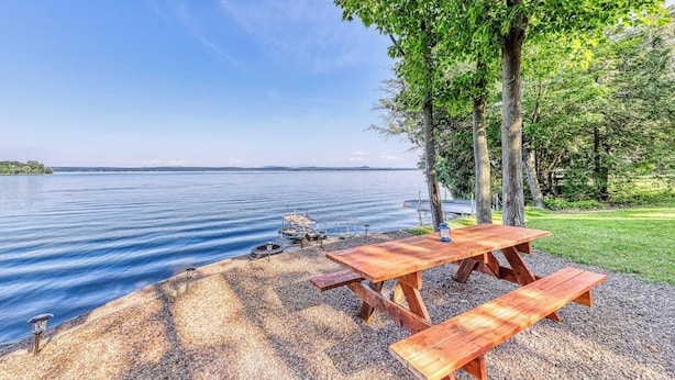 A lakeside picnic area with a wooden table and benches, surrounded by trees and a view of the water. - Dog-Friendly Lakefront Home with WiFi, Private Dock & Firepit (Grand Isle)