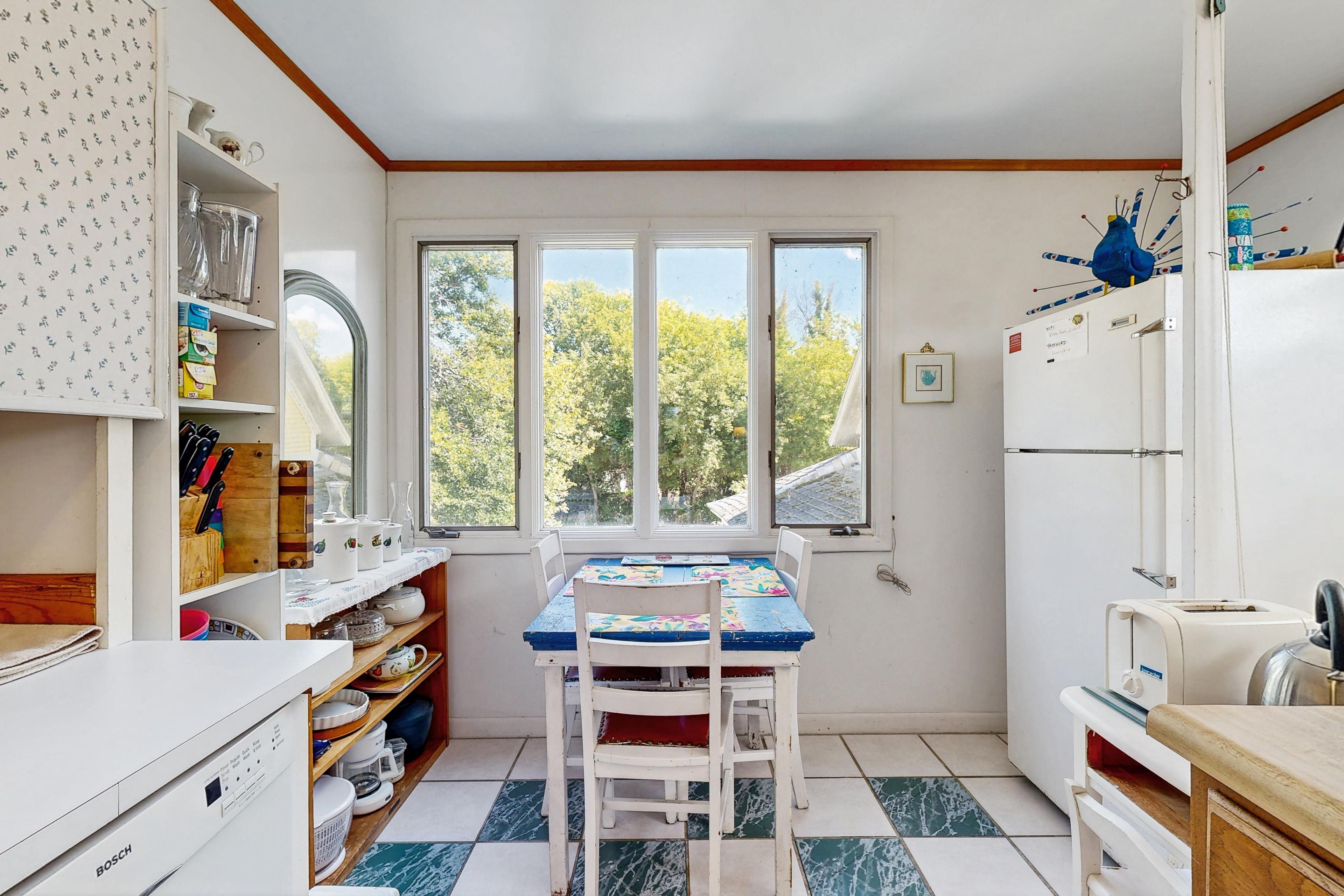 Una cocina con mesa, sillas, refrigerador y ventana con vistas a árboles.