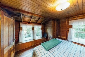 A wooden cabin room with a bed, a window with curtains, and a ceiling light.