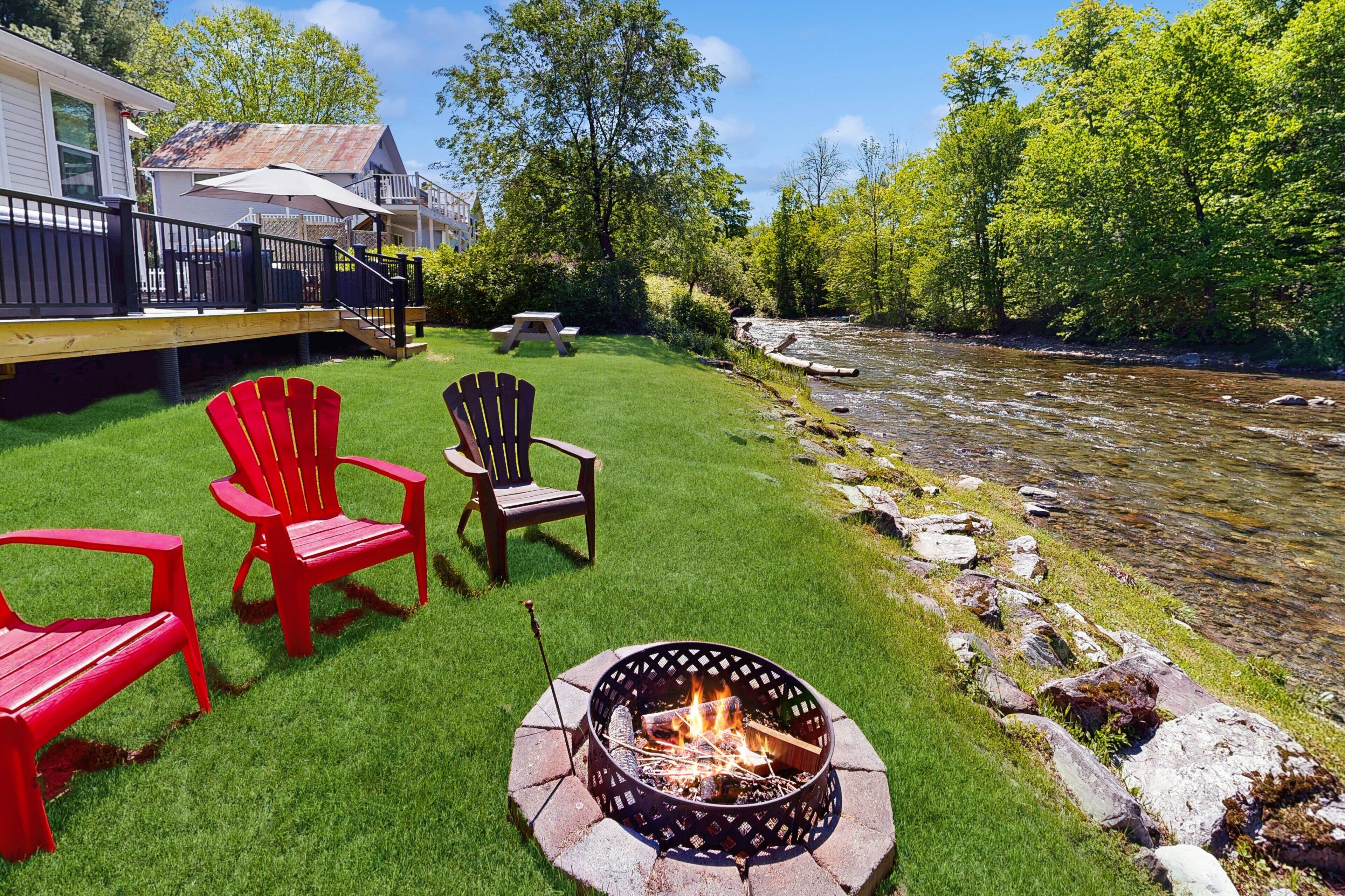Een achtertuin met een vuurkuil, rode Adirondack-stoelen en uitzicht op een rivier.