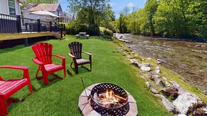 En trädgård med en eldstad, röda Adirondack-stolar och utsikt över en flod.