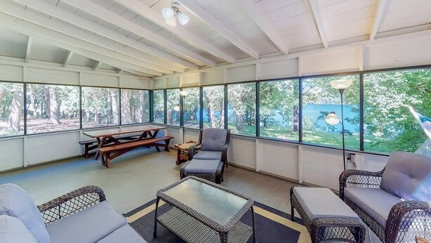 A covered outdoor seating area with wicker furniture, a picnic table, and large windows offering a view of the outdoors.