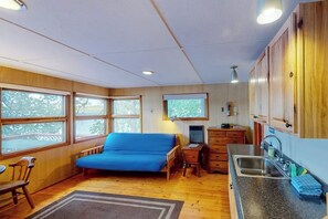 A compact room with a bed, a desk, a chair, a small kitchenette, and large windows.