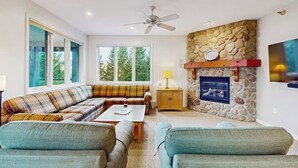 A living room with a stone fireplace, a ceiling fan, a sofa set, and a television mounted on the wall. - Spacious Mountain Retreat at Smugglers' Notch Resort w/ Great Views, Pool Access (Jeffersonville)