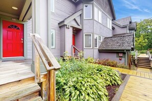 A two-story house with a red door, a small porch, and a wooden staircase leading to a deck.