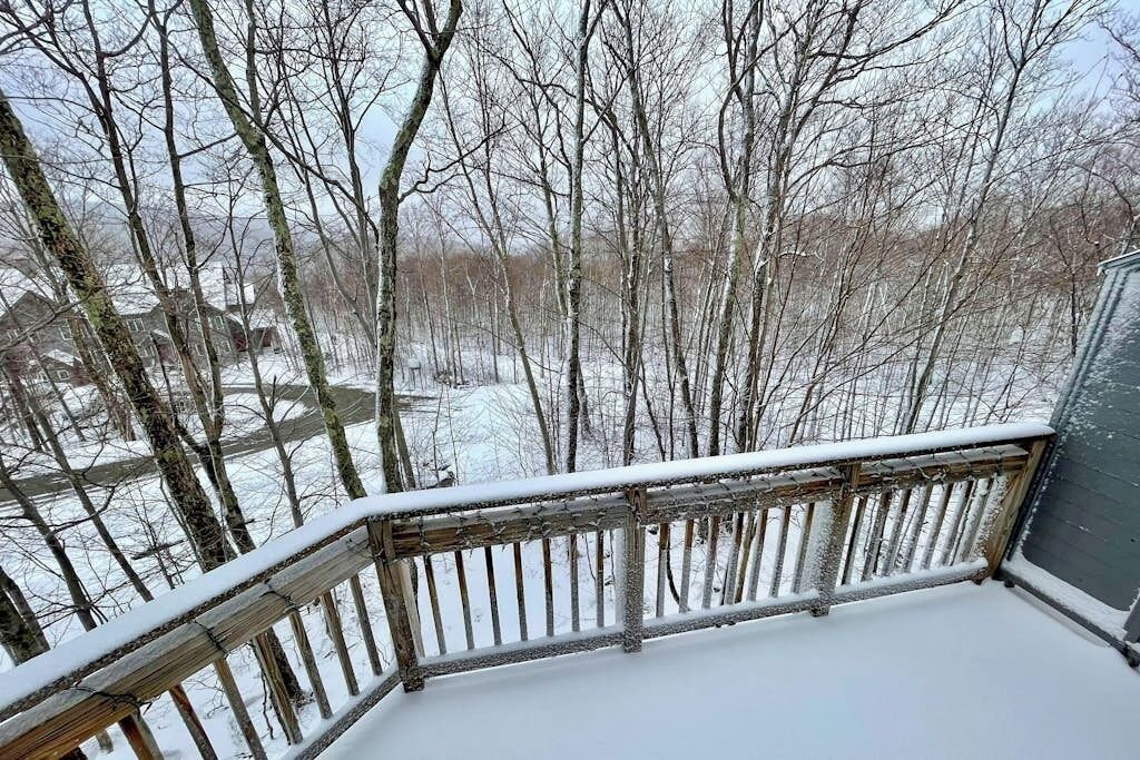 A snow-covered deck with a wooden railing, overlooking a forested area with bare trees.
