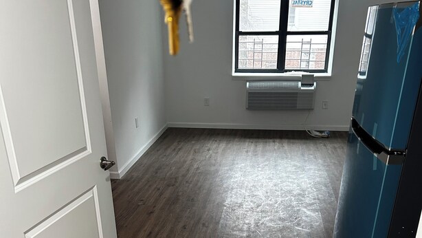 Una habitación con suelo de madera, una puerta blanca, un refrigerador azul y una ventana con persianas.