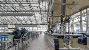 A spacious indoor area with high ceilings and large windows, featuring seating arrangements and service counters.