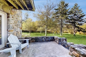 A stone exterior wall, a white chair, and a stone fire pit area with a grassy yard and trees in the background. - Mountain view house w/ two fireplaces, BBQ, w/d, jacuzzi, wifi, a/c (Elmore)