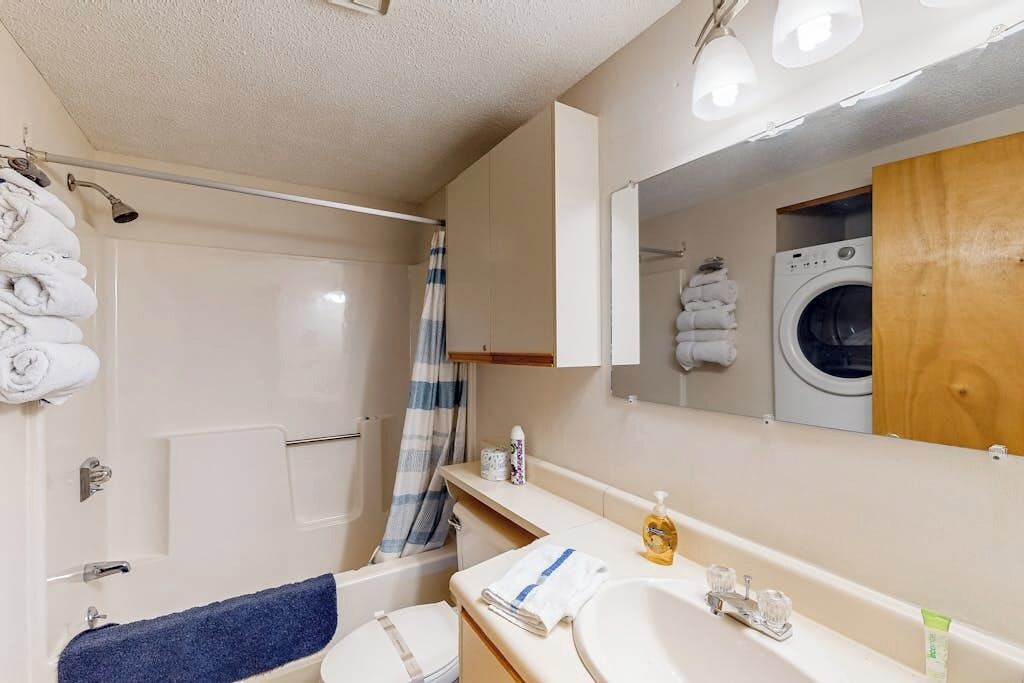 A bathroom with a shower, a toilet, a sink, a mirror, and a washing machine.
