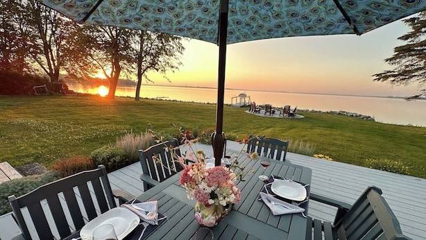 A dining table set outdoors with chairs, a table umbrella, and a view of the sunset over a body of water.