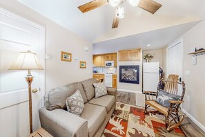 A living room with a sofa, a rocking chair, a lamp, a ceiling fan, and a kitchen area in the background.
